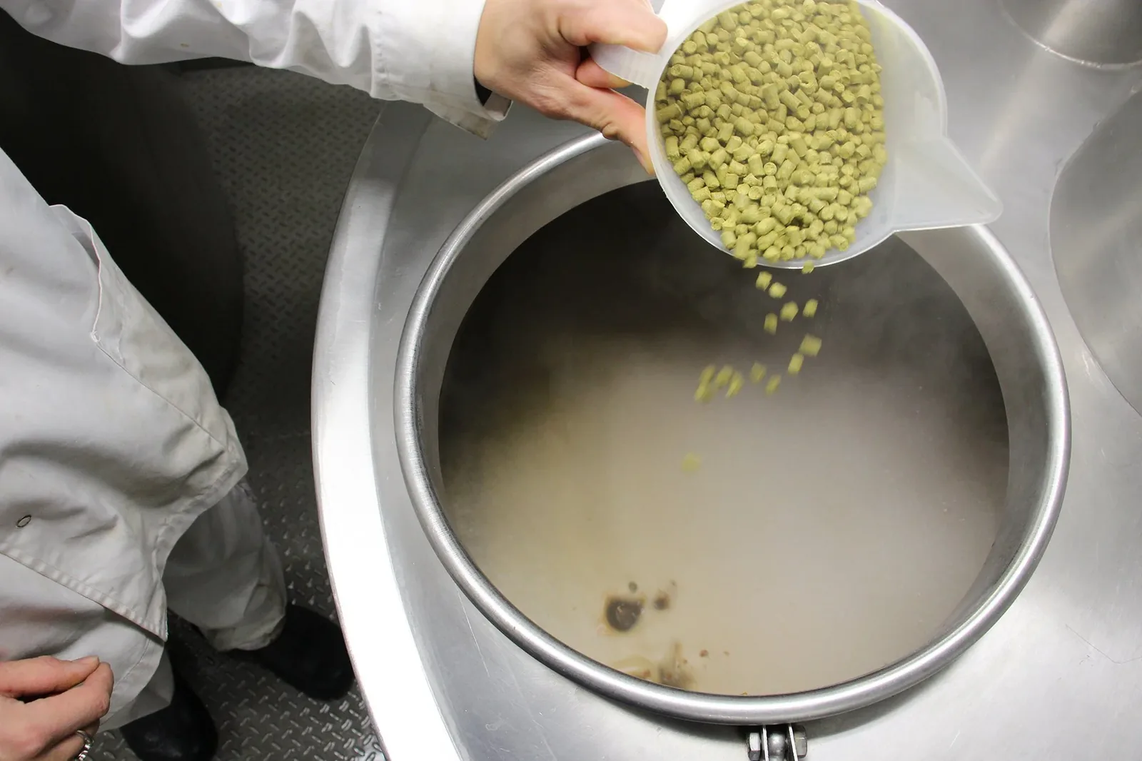 Mano vertiendo pellets de lúpulo desde un vaso medidor sobre el mosto en cocción